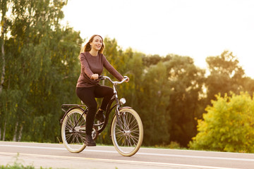 Obraz na płótnie Canvas Young attractive woman riding through the park after work. Beautiful lady cycling during sunset. Bike as a trendy transport. Healthy outdoors activity on a warm summer day. Bicycle trend in the city.