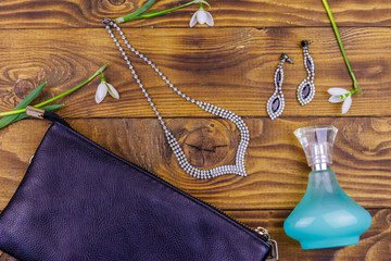 Women accessories on wooden background. Clutch bag, bottle of perfume, necklace, earrings and snowdrops on wood table. Beauty and fashion composition. Top view, flat lay