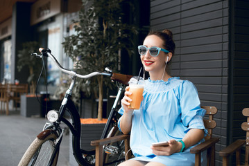 Fototapeta na wymiar Young beautiful lady drinking fresh smoothies on a sunny summer day during her ride. Riding a bicycle in the city. Bicycle tourism. Refreshing fruit drink. Active rest. Hipster on a bike. 