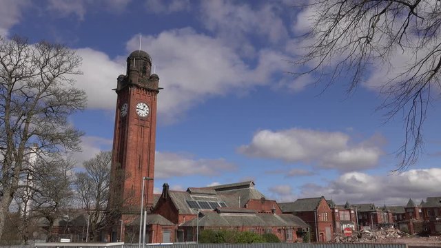Fast motion clouds in a bright blue sky passing the red brick Victorian clock tower of Stobhill Hospital. In the background are buildings in the process of being demolished.