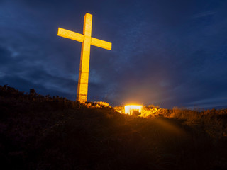 Cross in a field lit by a lamp, dark blue sky with clouds. Concept: how important is religion to people. How much they can do, devote and spend.