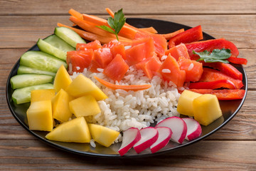 Top view of a Poke salad from hawaii in black plate on wooden background. Close up. Soft focus. To prepare it just take rice, salmon and any vegetables.