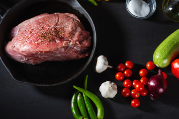 Raw fresh beef in a frying pan and vegetables. Preparation for roast beef. Black wooden table background. Close up. Selective focus.