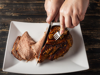 Female Chef cutting roast beef on white plate, hands, close up. Old, black wooden table.