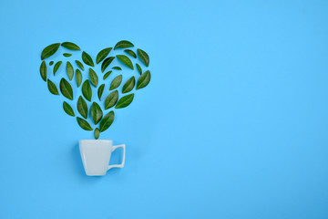 Coffee cup with Green leaves arranged in heart shape over blue background. Love and morning concept. Flat lay, top view, copy space