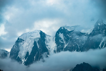 Low cloud before huge glacier. Giant snowy rocky mountains under cloudy sky. Thick fog in mountains at early morning. Impenetrable fog. Cold rocks. Dark atmospheric landscape. Tranquil atmosphere.