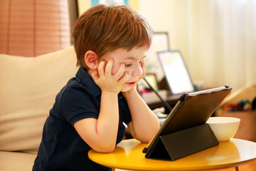 Child using tablet pc on bed at home. Cute boy on sofa is watching cartoon, playing games and learning from laptop. Education, fun, leisure, happiness, modern computer technology and communication.