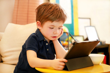 Child using tablet pc on bed at home. Cute boy on sofa is watching cartoon, playing games and learning from laptop. Education, fun, leisure, happiness, modern computer technology and communication.