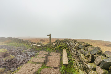 A view of a hill summit point area with wooden trail path sign and bench along a stone wall with walker wooden gate under misty cloud sky