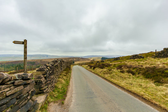 A view of a rural road lane with stone wall on the side, walking path sign and green vegetation under a white cloudy sky