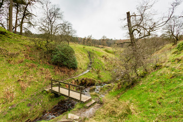 A view of a wooden bridge over a stream with green slopes under a white cloudy sky
