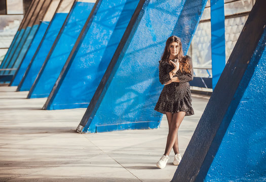 Lonely beautiful girl standing between blue columns of grunge city area. Young woman drinking coffee in urban sunny space