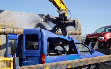 Claw from a crane at junkyard left an old blue car at wrecker press to get it into cube for...