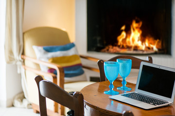 Laptop standing on ancient, vintage and old oak table in cozy living room next to fireplace with glowing, soft burning fire. Empty glasses of wine or beer. Waiting guests. Concept of working at home
