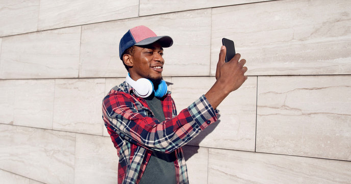 Portrait stylish smiling african man taking selfie picture by phone with headphones in baseball cap, plaid shirt on city street over gray wall background