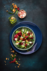 Beet or beetroot salad with fresh arugula, radicchio, soft cheese and walnuts on plate with fork, dressing and spices on blue kitchen table background, copy space, top view