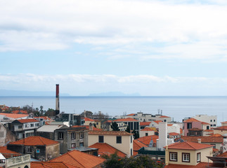 high coastal view of houses with red tiled roofs and old factory chimney in the city of funchal with a blue sky with white clouds over the sea