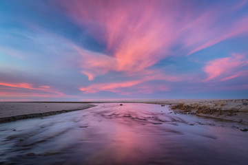 Pink clouds and blue sky over the fast river on the sandy beach at sunset
