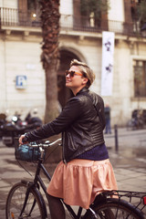 A young attractive woman is sitting on a bike and smiling. A blond female in a leather jacket and pink dress and sunglasses on Barcelona street on a sunny day. Bicycle ride. 