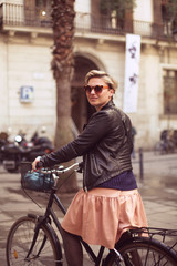 A young attractive woman is sitting on a bike and smiling. A blond female in a leather jacket and pink dress and sunglasses on Barcelona street on a sunny day. Bicycle ride. 