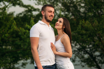 Young couple in white stand on the hill. Background of trees and lake