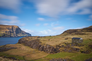 Faroese grazing or pasture land for pasture sheep with traditional old stone herdsman house on top of high cliff in great fjord of north atlantic ocean in sunny day.