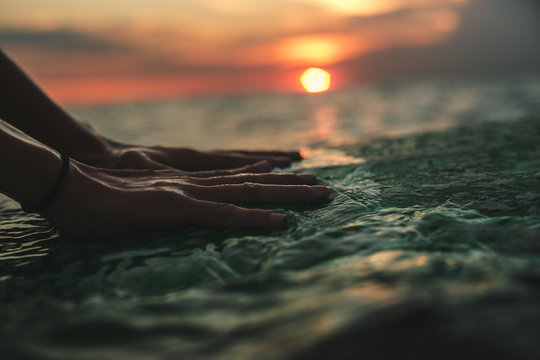 Close-up of woman's hands touching surface of surfboard in the water, beautiful sunset on background.