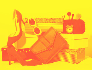 Top view female outfit composition: accessories shoes, handbag clutch, sunglasses, cosmetics makeup, alarm clock on carton boxes on red yellow background.