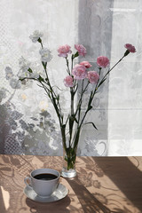 Flowers in a vase and a cup of coffee on a sunny window in a rustic house on a wooden table, a beautiful light background of early morning and good mood, long shadows from flowers and a window