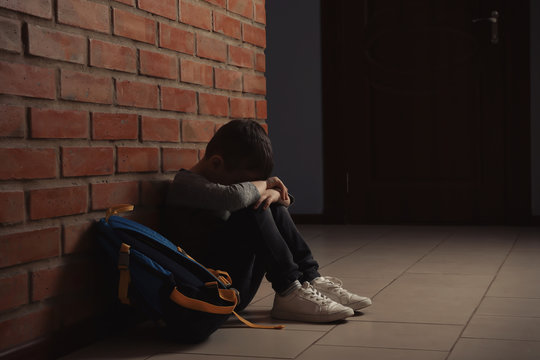 Sad little boy with backpack sitting on floor near brick wall indoors