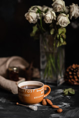 Big orange mug of black tea accompanied with white fade roses in glass vase, almonds, cones, small jam jar and sugar bar on dark background