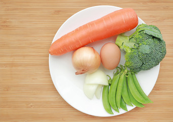 Fresh vegetable in white plate against wood board background (broccoli, carrot, egg, onion and sweet pea)