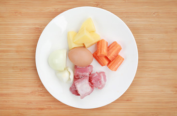 Carrot, egg, onion, pork bone and potato on white plate against wooden background Ingredients for boiling soup.