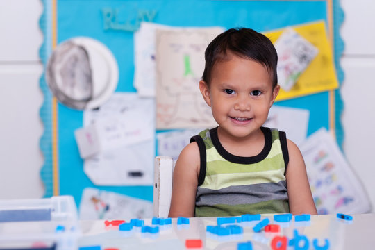 A cute hispanic boy sitting in daycare with alphabet letters on the table and a colorful bulletin board in the background.