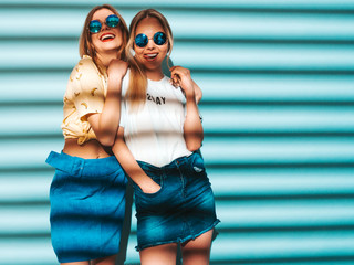 Two young beautiful smiling blond hipster girls in trendy summer colorful T-shirt clothes. Sexy carefree women posing near blue wall in round sunglasses. Positive models having fun