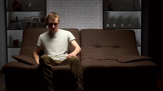young man sits down on a sofa and turns on the TV using a remote control.