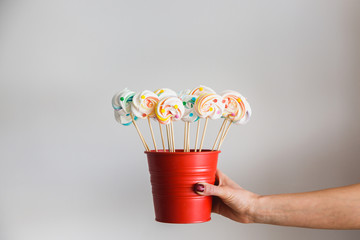 Hand holding colorful meringue lollipops on a stick in red pail, white background.