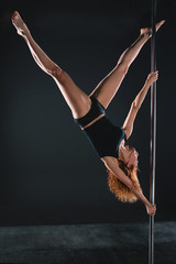 Fototapeta na wymiar Pole sports: beautiful young athletic fit strong redhead woman gymnast in black T-shirt and shorts doing sport exercises (training, work out) on the pole on dark background