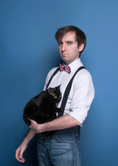 handsome man in shirt, suspender and pink bow tie holding cute black cat and looking at camera on blue background with copy space