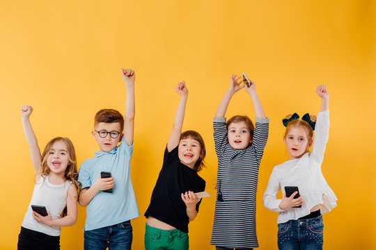 5 beautiful children use smartphone, raise hands up, happy, small glasses in glasses and 4 little girls, isolated yellow background, copy space