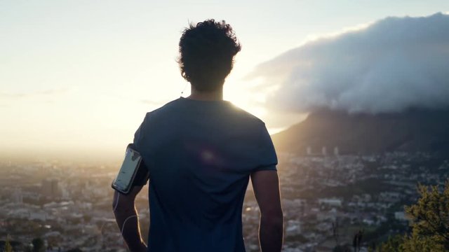 Shot of a young white male facing backwards at the camera and overlooking the view of the city and nature at sunrise