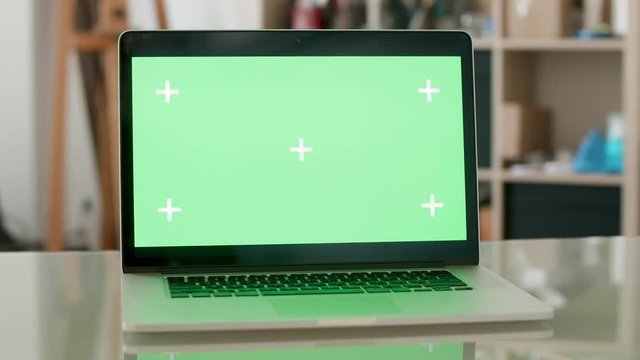 Parallax shot of a laptop with green scren on and people on the background. Designers office concept. Just notebook on a desk.