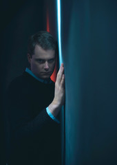 Suspicious man in black sweater stands against wall in red and blue light.