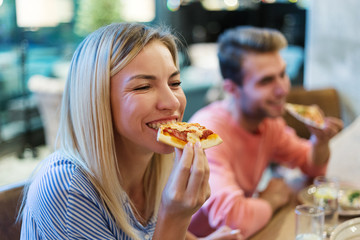 Joyful blonde woman enjoying eating delicious pizza at dinner in Italian restaurant, her male...