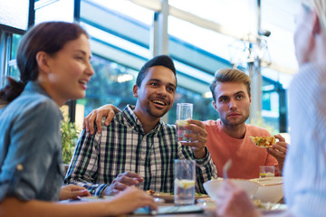 Young multi-ethnic friends drinking lemonade, eating pizza and listening to story being told by their girlfriend during dinner party at restaurant