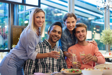Portrait of four happy young friends posing for photo together at dining table, smiling at camera and toasting with lemonade in restaurant