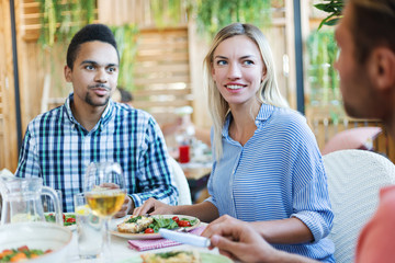 Young Caucasian woman and Latin American man listening to their unrecognizable friend at dinner