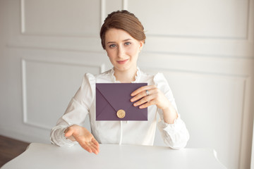 girl in a white business style shirt with a classic hairdo on brunette hair holding a welcome envelope in her hand sitting at a table