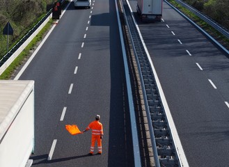 A highway worker with a high visibility work suit waves the orange flag to slow down trucks traffic before the roadblock