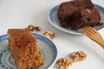 Top view of sponge cake with carrots and brownie with nuts and wooden fork on blue and white plate. On white wooden background